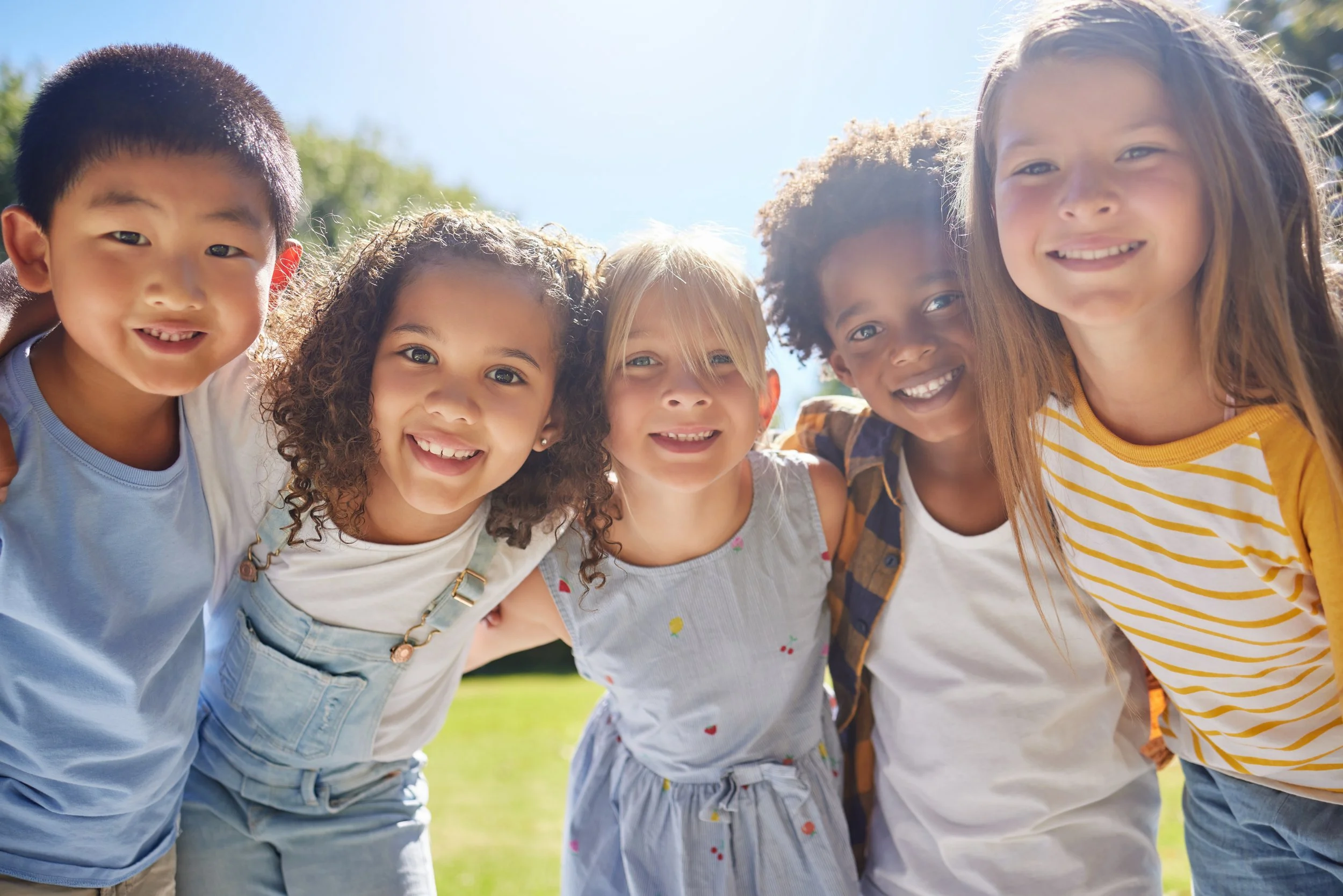 Five smiling kids with their arms around each other outdoors on a sunny day